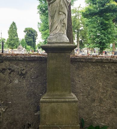 Zdjęcie nr 2: Nagrobek wolnostojący na rozbudowanym cokole, zwieńczony figurą Matki Boskiej. Cokół jest gładki, wieloczęściowy, z której część niższa jest szersza od górnej. Dolna jest prostopadłościenna, z uskokiem, na górnej części na frontowej ścianie inskrypcja. Figura Matki Boskiej w typie Niepokalanej ukazuje Marię w całej postaci, stojącą frontalnie, z rękami złożonymi do modlitwy i półksiężycem pod prawą stopą. Ma owalną, zamyśloną twarz o łagodnych rysach, oczy przymknięte, nos prosty, usta wąskie. Jest ubrana w długą, przewiązaną w pasie suknię i siegający ziemi płaszcz, który osłania jej głowę, plecy i jest założony za pasek przy lewym biodrze. 