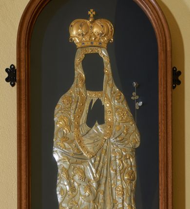 Zdjęcie nr 1: Sukienka na obraz Matki Boskiej umieszczona w przeszklonej oprawie w kształcie stojącego prostokąta, zamkniętego łukiem półkolistym. Suknia Matki Boskiej z długimi rękawami ułożonymi na piersi w geście modlitwy, u szyi dekorowana kaboszonami i rautami. Płaszcz założony na głowę, ciasno oplatający sylwetkę, zdobiony ulistnioną wicią z owocami i kwiatami granatu. Na brzegach jest dekorowany ozdobnym pasem z rautami i kaboszonami. U dołu półksiężyc, a na głowie zamknięta korona z krzyżykiem, wysadzana kamieniami. Z prawej strony zawieszona srebrna róża. Półksiężyc, korona i dekoracja roślinna złocone. Rama drewniana, profilowana i malowana w kolorze brązu. Tło ciemnoniebieskie.