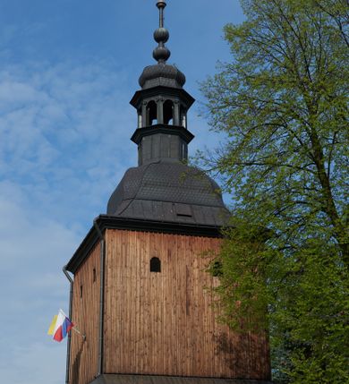 Zdjęcie nr 1: Dzwonnica na planie kwadratu, dwukondygnacyjna, zwieńczona hełmem z latarnią na szczycie. Kondygnacja dolna, murowana, na cokole, przepruta przez środek bramą o półkolistym wykroju, ujęta w narożnikach lizenami, podtrzymującymi gzyms i nakryta pulpitowym daszkiem. Kondygnacja górna, drewniana, węższa i wyższa, oszalowana deskami w układzie pionowym z listwowaniem, z dwoma małymi otworami okiennymi na każdej ścianie. Hełm cebulasty z ośmioboczną latarnią, zwieńczoną zwielokrotnioną formą spłaszczonej kopułki z kulą i ażurowym krzyżem na szczycie. Trzy dzwony zawieszone na oddzielnej konstrukcji w kształcie trzech krosien. Przyziemie oraz klatka schodowa prowadząca na dzwony nakryte ceglanymi sklepieniami kolebkowymi, a ponad sklepieniem dolnej kondygnacji strop drewniany nakryty powałą. Schody zewnętrzne, prowadzące do bramy pokryte okładziną kamienną, z jednej strony betonowe. 
