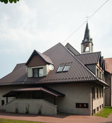 Zdjęcie nr 10: Kościół Podwyższenia Krzyża Świętego w Janowicach jest zlokalizowany na niewielkim wzniesieniu, w centrum prostokątnego placu otoczonego murowanym ogrodzeniem podzielonym na przęsła. W południowo-zachodnim narożu terenu przykościelnego usytuowana jest parawanowa dzwonnica. Kościół to budowla murowana z cegły, jednonawowa na planie krzyża łacińskiego. Do czteroprzęsłowej nawy przylega wyniesione na czterostopniowym podwyższeniu prezbiterium zamknięte prostą ścianą. Do prezbiterium od zachodu przylega zakrystia na planie zbliżonym do litery „L”, od wschodu aneks na analogicznym planie. Zakrystia skomunikowana jest z dobudowaną od południa dwukondygnacyjną i podpiwniczoną  plebanią. Wnętrze świątyni przekryte jest płaskim stropem o konstrukcji żelbetowej. Ściany w dolnej części artykułowane są za pomocą boazerii, w wyższych partiach podziały kontynuowane są w polichromii. W górnych partiach ścian (poza prezbiterium) znajdują się ukośne sufity, w nawie dodatkowo poprzedzone profilowanym gzymsem. W najwyższym rzędzie płycin mieszczą się płaskorzeźbione, stylizowane rozety z motywami geometrycznymi; analogiczne rozety zdobią parapet chóru muzycznego. Nawę doświetlają okna w kształcie stojącego prostokąta zamkniętego półkoliście: po jednym w przęsłach od drugiego do czwartego i jedno nieco większe, ponad chórem w ścianie północnej. W ścianach zamykających transept znajdują się po trzy duże otwory okienne złożone z umieszczonego po środku dużego okna w kształcie odwróconej podkowy oraz flankujących go mniejszych okien w kształcie stojącego prostokąta zamkniętego półkoliście. Główne wejście do świątyni znajduje się na osi budynku, w ścianie północnej, wejście boczne usytuowane jest w północnej ścianie zachodniego ramienia transeptu. Na szerokości pierwszego od północy przęsła nawy znajduje się chór muzyczny o konstrukcji żelbetowej, wsparty na dwóch filarach. Pod nim mieści się kruchta. Parapet chóru muzycznego jest murowany, prosty, w części środkowej trójbocznie wyłamany do wnętrza kościoła. W osi środkowej trójdzielnej fasady kościoła umieszczona wieża na planie kwadratu, nieco cofnięta w stosunku do lica elewacji, którą ujmują ukośnie ustawione ściany. Wejście do świątyni znajduje się na osi fasady. Jest zadaszone płaskim dachem; prowadzą do niego siedmiostopniowe schody. Od strony zachodniej zlokalizowany jest podjazd dla niepełnosprawnych. Otwór wejściowy ma kształt prostokąta, wewnątrz znajdują się drewniane, dwuskrzydłowe drzwi. Powyżej umiejscowione jest duże okno w kształcie stojącego prostokąta zamkniętego półkoliście. W górnej części wieży umieszczono trzy wysmukłe okna zamknięte półkoliście. W zwieńczeniu mieści się ażurowy prześwit pełniący funkcję tarasu widokowego. Wieżę wieńczy hełm w kształcie wysmukłego ostrosłupa z krzyżem. Elewacje boczne pozbawione są dekoracji.


