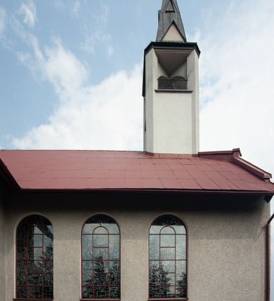 Zdjęcie nr 11: Kościół Podwyższenia Krzyża Świętego w Janowicach jest zlokalizowany na niewielkim wzniesieniu, w centrum prostokątnego placu otoczonego murowanym ogrodzeniem podzielonym na przęsła. W południowo-zachodnim narożu terenu przykościelnego usytuowana jest parawanowa dzwonnica. Kościół to budowla murowana z cegły, jednonawowa na planie krzyża łacińskiego. Do czteroprzęsłowej nawy przylega wyniesione na czterostopniowym podwyższeniu prezbiterium zamknięte prostą ścianą. Do prezbiterium od zachodu przylega zakrystia na planie zbliżonym do litery „L”, od wschodu aneks na analogicznym planie. Zakrystia skomunikowana jest z dobudowaną od południa dwukondygnacyjną i podpiwniczoną  plebanią. Wnętrze świątyni przekryte jest płaskim stropem o konstrukcji żelbetowej. Ściany w dolnej części artykułowane są za pomocą boazerii, w wyższych partiach podziały kontynuowane są w polichromii. W górnych partiach ścian (poza prezbiterium) znajdują się ukośne sufity, w nawie dodatkowo poprzedzone profilowanym gzymsem. W najwyższym rzędzie płycin mieszczą się płaskorzeźbione, stylizowane rozety z motywami geometrycznymi; analogiczne rozety zdobią parapet chóru muzycznego. Nawę doświetlają okna w kształcie stojącego prostokąta zamkniętego półkoliście: po jednym w przęsłach od drugiego do czwartego i jedno nieco większe, ponad chórem w ścianie północnej. W ścianach zamykających transept znajdują się po trzy duże otwory okienne złożone z umieszczonego po środku dużego okna w kształcie odwróconej podkowy oraz flankujących go mniejszych okien w kształcie stojącego prostokąta zamkniętego półkoliście. Główne wejście do świątyni znajduje się na osi budynku, w ścianie północnej, wejście boczne usytuowane jest w północnej ścianie zachodniego ramienia transeptu. Na szerokości pierwszego od północy przęsła nawy znajduje się chór muzyczny o konstrukcji żelbetowej, wsparty na dwóch filarach. Pod nim mieści się kruchta. Parapet chóru muzycznego jest murowany, prosty, w części środkowej trójbocznie wyłamany do wnętrza kościoła. W osi środkowej trójdzielnej fasady kościoła umieszczona wieża na planie kwadratu, nieco cofnięta w stosunku do lica elewacji, którą ujmują ukośnie ustawione ściany. Wejście do świątyni znajduje się na osi fasady. Jest zadaszone płaskim dachem; prowadzą do niego siedmiostopniowe schody. Od strony zachodniej zlokalizowany jest podjazd dla niepełnosprawnych. Otwór wejściowy ma kształt prostokąta, wewnątrz znajdują się drewniane, dwuskrzydłowe drzwi. Powyżej umiejscowione jest duże okno w kształcie stojącego prostokąta zamkniętego półkoliście. W górnej części wieży umieszczono trzy wysmukłe okna zamknięte półkoliście. W zwieńczeniu mieści się ażurowy prześwit pełniący funkcję tarasu widokowego. Wieżę wieńczy hełm w kształcie wysmukłego ostrosłupa z krzyżem. Elewacje boczne pozbawione są dekoracji.


