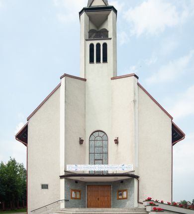 Zdjęcie nr 1: Kościół Podwyższenia Krzyża Świętego w Janowicach jest zlokalizowany na niewielkim wzniesieniu, w centrum prostokątnego placu otoczonego murowanym ogrodzeniem podzielonym na przęsła. W południowo-zachodnim narożu terenu przykościelnego usytuowana jest parawanowa dzwonnica. Kościół to budowla murowana z cegły, jednonawowa na planie krzyża łacińskiego. Do czteroprzęsłowej nawy przylega wyniesione na czterostopniowym podwyższeniu prezbiterium zamknięte prostą ścianą. Do prezbiterium od zachodu przylega zakrystia na planie zbliżonym do litery „L”, od wschodu aneks na analogicznym planie. Zakrystia skomunikowana jest z dobudowaną od południa dwukondygnacyjną i podpiwniczoną  plebanią. Wnętrze świątyni przekryte jest płaskim stropem o konstrukcji żelbetowej. Ściany w dolnej części artykułowane są za pomocą boazerii, w wyższych partiach podziały kontynuowane są w polichromii. W górnych partiach ścian (poza prezbiterium) znajdują się ukośne sufity, w nawie dodatkowo poprzedzone profilowanym gzymsem. W najwyższym rzędzie płycin mieszczą się płaskorzeźbione, stylizowane rozety z motywami geometrycznymi; analogiczne rozety zdobią parapet chóru muzycznego. Nawę doświetlają okna w kształcie stojącego prostokąta zamkniętego półkoliście: po jednym w przęsłach od drugiego do czwartego i jedno nieco większe, ponad chórem w ścianie północnej. W ścianach zamykających transept znajdują się po trzy duże otwory okienne złożone z umieszczonego po środku dużego okna w kształcie odwróconej podkowy oraz flankujących go mniejszych okien w kształcie stojącego prostokąta zamkniętego półkoliście. Główne wejście do świątyni znajduje się na osi budynku, w ścianie północnej, wejście boczne usytuowane jest w północnej ścianie zachodniego ramienia transeptu. Na szerokości pierwszego od północy przęsła nawy znajduje się chór muzyczny o konstrukcji żelbetowej, wsparty na dwóch filarach. Pod nim mieści się kruchta. Parapet chóru muzycznego jest murowany, prosty, w części środkowej trójbocznie wyłamany do wnętrza kościoła. W osi środkowej trójdzielnej fasady kościoła umieszczona wieża na planie kwadratu, nieco cofnięta w stosunku do lica elewacji, którą ujmują ukośnie ustawione ściany. Wejście do świątyni znajduje się na osi fasady. Jest zadaszone płaskim dachem; prowadzą do niego siedmiostopniowe schody. Od strony zachodniej zlokalizowany jest podjazd dla niepełnosprawnych. Otwór wejściowy ma kształt prostokąta, wewnątrz znajdują się drewniane, dwuskrzydłowe drzwi. Powyżej umiejscowione jest duże okno w kształcie stojącego prostokąta zamkniętego półkoliście. W górnej części wieży umieszczono trzy wysmukłe okna zamknięte półkoliście. W zwieńczeniu mieści się ażurowy prześwit pełniący funkcję tarasu widokowego. Wieżę wieńczy hełm w kształcie wysmukłego ostrosłupa z krzyżem. Elewacje boczne pozbawione są dekoracji.


