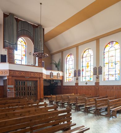 Zdjęcie nr 7: Kościół Podwyższenia Krzyża Świętego w Janowicach jest zlokalizowany na niewielkim wzniesieniu, w centrum prostokątnego placu otoczonego murowanym ogrodzeniem podzielonym na przęsła. W południowo-zachodnim narożu terenu przykościelnego usytuowana jest parawanowa dzwonnica. Kościół to budowla murowana z cegły, jednonawowa na planie krzyża łacińskiego. Do czteroprzęsłowej nawy przylega wyniesione na czterostopniowym podwyższeniu prezbiterium zamknięte prostą ścianą. Do prezbiterium od zachodu przylega zakrystia na planie zbliżonym do litery „L”, od wschodu aneks na analogicznym planie. Zakrystia skomunikowana jest z dobudowaną od południa dwukondygnacyjną i podpiwniczoną  plebanią. Wnętrze świątyni przekryte jest płaskim stropem o konstrukcji żelbetowej. Ściany w dolnej części artykułowane są za pomocą boazerii, w wyższych partiach podziały kontynuowane są w polichromii. W górnych partiach ścian (poza prezbiterium) znajdują się ukośne sufity, w nawie dodatkowo poprzedzone profilowanym gzymsem. W najwyższym rzędzie płycin mieszczą się płaskorzeźbione, stylizowane rozety z motywami geometrycznymi; analogiczne rozety zdobią parapet chóru muzycznego. Nawę doświetlają okna w kształcie stojącego prostokąta zamkniętego półkoliście: po jednym w przęsłach od drugiego do czwartego i jedno nieco większe, ponad chórem w ścianie północnej. W ścianach zamykających transept znajdują się po trzy duże otwory okienne złożone z umieszczonego po środku dużego okna w kształcie odwróconej podkowy oraz flankujących go mniejszych okien w kształcie stojącego prostokąta zamkniętego półkoliście. Główne wejście do świątyni znajduje się na osi budynku, w ścianie północnej, wejście boczne usytuowane jest w północnej ścianie zachodniego ramienia transeptu. Na szerokości pierwszego od północy przęsła nawy znajduje się chór muzyczny o konstrukcji żelbetowej, wsparty na dwóch filarach. Pod nim mieści się kruchta. Parapet chóru muzycznego jest murowany, prosty, w części środkowej trójbocznie wyłamany do wnętrza kościoła. W osi środkowej trójdzielnej fasady kościoła umieszczona wieża na planie kwadratu, nieco cofnięta w stosunku do lica elewacji, którą ujmują ukośnie ustawione ściany. Wejście do świątyni znajduje się na osi fasady. Jest zadaszone płaskim dachem; prowadzą do niego siedmiostopniowe schody. Od strony zachodniej zlokalizowany jest podjazd dla niepełnosprawnych. Otwór wejściowy ma kształt prostokąta, wewnątrz znajdują się drewniane, dwuskrzydłowe drzwi. Powyżej umiejscowione jest duże okno w kształcie stojącego prostokąta zamkniętego półkoliście. W górnej części wieży umieszczono trzy wysmukłe okna zamknięte półkoliście. W zwieńczeniu mieści się ażurowy prześwit pełniący funkcję tarasu widokowego. Wieżę wieńczy hełm w kształcie wysmukłego ostrosłupa z krzyżem. Elewacje boczne pozbawione są dekoracji.


