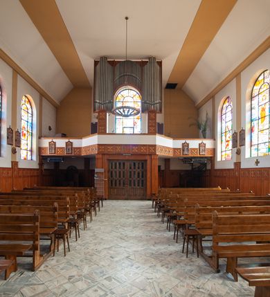 Zdjęcie nr 5: Kościół Podwyższenia Krzyża Świętego w Janowicach jest zlokalizowany na niewielkim wzniesieniu, w centrum prostokątnego placu otoczonego murowanym ogrodzeniem podzielonym na przęsła. W południowo-zachodnim narożu terenu przykościelnego usytuowana jest parawanowa dzwonnica. Kościół to budowla murowana z cegły, jednonawowa na planie krzyża łacińskiego. Do czteroprzęsłowej nawy przylega wyniesione na czterostopniowym podwyższeniu prezbiterium zamknięte prostą ścianą. Do prezbiterium od zachodu przylega zakrystia na planie zbliżonym do litery „L”, od wschodu aneks na analogicznym planie. Zakrystia skomunikowana jest z dobudowaną od południa dwukondygnacyjną i podpiwniczoną  plebanią. Wnętrze świątyni przekryte jest płaskim stropem o konstrukcji żelbetowej. Ściany w dolnej części artykułowane są za pomocą boazerii, w wyższych partiach podziały kontynuowane są w polichromii. W górnych partiach ścian (poza prezbiterium) znajdują się ukośne sufity, w nawie dodatkowo poprzedzone profilowanym gzymsem. W najwyższym rzędzie płycin mieszczą się płaskorzeźbione, stylizowane rozety z motywami geometrycznymi; analogiczne rozety zdobią parapet chóru muzycznego. Nawę doświetlają okna w kształcie stojącego prostokąta zamkniętego półkoliście: po jednym w przęsłach od drugiego do czwartego i jedno nieco większe, ponad chórem w ścianie północnej. W ścianach zamykających transept znajdują się po trzy duże otwory okienne złożone z umieszczonego po środku dużego okna w kształcie odwróconej podkowy oraz flankujących go mniejszych okien w kształcie stojącego prostokąta zamkniętego półkoliście. Główne wejście do świątyni znajduje się na osi budynku, w ścianie północnej, wejście boczne usytuowane jest w północnej ścianie zachodniego ramienia transeptu. Na szerokości pierwszego od północy przęsła nawy znajduje się chór muzyczny o konstrukcji żelbetowej, wsparty na dwóch filarach. Pod nim mieści się kruchta. Parapet chóru muzycznego jest murowany, prosty, w części środkowej trójbocznie wyłamany do wnętrza kościoła. W osi środkowej trójdzielnej fasady kościoła umieszczona wieża na planie kwadratu, nieco cofnięta w stosunku do lica elewacji, którą ujmują ukośnie ustawione ściany. Wejście do świątyni znajduje się na osi fasady. Jest zadaszone płaskim dachem; prowadzą do niego siedmiostopniowe schody. Od strony zachodniej zlokalizowany jest podjazd dla niepełnosprawnych. Otwór wejściowy ma kształt prostokąta, wewnątrz znajdują się drewniane, dwuskrzydłowe drzwi. Powyżej umiejscowione jest duże okno w kształcie stojącego prostokąta zamkniętego półkoliście. W górnej części wieży umieszczono trzy wysmukłe okna zamknięte półkoliście. W zwieńczeniu mieści się ażurowy prześwit pełniący funkcję tarasu widokowego. Wieżę wieńczy hełm w kształcie wysmukłego ostrosłupa z krzyżem. Elewacje boczne pozbawione są dekoracji.


