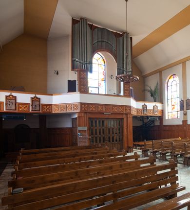 Zdjęcie nr 6: Kościół Podwyższenia Krzyża Świętego w Janowicach jest zlokalizowany na niewielkim wzniesieniu, w centrum prostokątnego placu otoczonego murowanym ogrodzeniem podzielonym na przęsła. W południowo-zachodnim narożu terenu przykościelnego usytuowana jest parawanowa dzwonnica. Kościół to budowla murowana z cegły, jednonawowa na planie krzyża łacińskiego. Do czteroprzęsłowej nawy przylega wyniesione na czterostopniowym podwyższeniu prezbiterium zamknięte prostą ścianą. Do prezbiterium od zachodu przylega zakrystia na planie zbliżonym do litery „L”, od wschodu aneks na analogicznym planie. Zakrystia skomunikowana jest z dobudowaną od południa dwukondygnacyjną i podpiwniczoną  plebanią. Wnętrze świątyni przekryte jest płaskim stropem o konstrukcji żelbetowej. Ściany w dolnej części artykułowane są za pomocą boazerii, w wyższych partiach podziały kontynuowane są w polichromii. W górnych partiach ścian (poza prezbiterium) znajdują się ukośne sufity, w nawie dodatkowo poprzedzone profilowanym gzymsem. W najwyższym rzędzie płycin mieszczą się płaskorzeźbione, stylizowane rozety z motywami geometrycznymi; analogiczne rozety zdobią parapet chóru muzycznego. Nawę doświetlają okna w kształcie stojącego prostokąta zamkniętego półkoliście: po jednym w przęsłach od drugiego do czwartego i jedno nieco większe, ponad chórem w ścianie północnej. W ścianach zamykających transept znajdują się po trzy duże otwory okienne złożone z umieszczonego po środku dużego okna w kształcie odwróconej podkowy oraz flankujących go mniejszych okien w kształcie stojącego prostokąta zamkniętego półkoliście. Główne wejście do świątyni znajduje się na osi budynku, w ścianie północnej, wejście boczne usytuowane jest w północnej ścianie zachodniego ramienia transeptu. Na szerokości pierwszego od północy przęsła nawy znajduje się chór muzyczny o konstrukcji żelbetowej, wsparty na dwóch filarach. Pod nim mieści się kruchta. Parapet chóru muzycznego jest murowany, prosty, w części środkowej trójbocznie wyłamany do wnętrza kościoła. W osi środkowej trójdzielnej fasady kościoła umieszczona wieża na planie kwadratu, nieco cofnięta w stosunku do lica elewacji, którą ujmują ukośnie ustawione ściany. Wejście do świątyni znajduje się na osi fasady. Jest zadaszone płaskim dachem; prowadzą do niego siedmiostopniowe schody. Od strony zachodniej zlokalizowany jest podjazd dla niepełnosprawnych. Otwór wejściowy ma kształt prostokąta, wewnątrz znajdują się drewniane, dwuskrzydłowe drzwi. Powyżej umiejscowione jest duże okno w kształcie stojącego prostokąta zamkniętego półkoliście. W górnej części wieży umieszczono trzy wysmukłe okna zamknięte półkoliście. W zwieńczeniu mieści się ażurowy prześwit pełniący funkcję tarasu widokowego. Wieżę wieńczy hełm w kształcie wysmukłego ostrosłupa z krzyżem. Elewacje boczne pozbawione są dekoracji.


