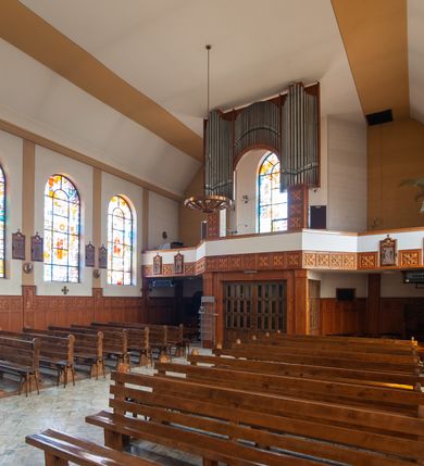 Zdjęcie nr 8: Kościół Podwyższenia Krzyża Świętego w Janowicach jest zlokalizowany na niewielkim wzniesieniu, w centrum prostokątnego placu otoczonego murowanym ogrodzeniem podzielonym na przęsła. W południowo-zachodnim narożu terenu przykościelnego usytuowana jest parawanowa dzwonnica. Kościół to budowla murowana z cegły, jednonawowa na planie krzyża łacińskiego. Do czteroprzęsłowej nawy przylega wyniesione na czterostopniowym podwyższeniu prezbiterium zamknięte prostą ścianą. Do prezbiterium od zachodu przylega zakrystia na planie zbliżonym do litery „L”, od wschodu aneks na analogicznym planie. Zakrystia skomunikowana jest z dobudowaną od południa dwukondygnacyjną i podpiwniczoną  plebanią. Wnętrze świątyni przekryte jest płaskim stropem o konstrukcji żelbetowej. Ściany w dolnej części artykułowane są za pomocą boazerii, w wyższych partiach podziały kontynuowane są w polichromii. W górnych partiach ścian (poza prezbiterium) znajdują się ukośne sufity, w nawie dodatkowo poprzedzone profilowanym gzymsem. W najwyższym rzędzie płycin mieszczą się płaskorzeźbione, stylizowane rozety z motywami geometrycznymi; analogiczne rozety zdobią parapet chóru muzycznego. Nawę doświetlają okna w kształcie stojącego prostokąta zamkniętego półkoliście: po jednym w przęsłach od drugiego do czwartego i jedno nieco większe, ponad chórem w ścianie północnej. W ścianach zamykających transept znajdują się po trzy duże otwory okienne złożone z umieszczonego po środku dużego okna w kształcie odwróconej podkowy oraz flankujących go mniejszych okien w kształcie stojącego prostokąta zamkniętego półkoliście. Główne wejście do świątyni znajduje się na osi budynku, w ścianie północnej, wejście boczne usytuowane jest w północnej ścianie zachodniego ramienia transeptu. Na szerokości pierwszego od północy przęsła nawy znajduje się chór muzyczny o konstrukcji żelbetowej, wsparty na dwóch filarach. Pod nim mieści się kruchta. Parapet chóru muzycznego jest murowany, prosty, w części środkowej trójbocznie wyłamany do wnętrza kościoła. W osi środkowej trójdzielnej fasady kościoła umieszczona wieża na planie kwadratu, nieco cofnięta w stosunku do lica elewacji, którą ujmują ukośnie ustawione ściany. Wejście do świątyni znajduje się na osi fasady. Jest zadaszone płaskim dachem; prowadzą do niego siedmiostopniowe schody. Od strony zachodniej zlokalizowany jest podjazd dla niepełnosprawnych. Otwór wejściowy ma kształt prostokąta, wewnątrz znajdują się drewniane, dwuskrzydłowe drzwi. Powyżej umiejscowione jest duże okno w kształcie stojącego prostokąta zamkniętego półkoliście. W górnej części wieży umieszczono trzy wysmukłe okna zamknięte półkoliście. W zwieńczeniu mieści się ażurowy prześwit pełniący funkcję tarasu widokowego. Wieżę wieńczy hełm w kształcie wysmukłego ostrosłupa z krzyżem. Elewacje boczne pozbawione są dekoracji.


