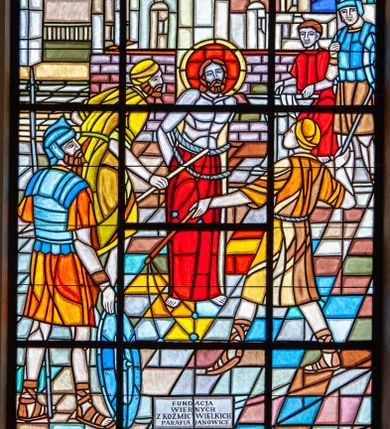 Zdjęcie nr 1: Witraż barwny w kształcie stojącego prostokąta zamkniętego półkoliście. W centralnej części wielopostaciowa kompozycja umieszczona na tle rozbudowanej architektury przedstawiająca biczowanie Chrystusa. Chrystus ukazany w pozycji stojącej z ciałem zwróconym w trzech czwartych w lewo; skrępowany sznurem przy kolumnie biczowania. Tors nagi, wokół pasa udrapowana czerwona, sięgająca ziemi tkanina. Wokół głowy Chrystusa czerwony nimb z żółtym obramowaniem. Postać Jezusa otaczają oprawcy ubrani w krótkie pomarańczowo-żółte tuniki oraz nakrycia głowy w tych samych kolorach ukazani w dynamicznych pozach, trzymający rózgi (bicze). Z lewej strony kompozycji stojąca tyłem, zwrócona w trzech czwartych w prawo postać rzymskiego żołnierza z włócznią i tarczą. Na drugim planie, z prawej strony witraża ukazani kolejny żołnierz oraz Poncjusz Piłat, umywający ręce w kamiennej misie przedstawiony w długiej, czerwonej szacie. Trzeci plan stanowi rozbudowane tło architektoniczne oraz wielobarwne niebo (w kolorach: czerwonym, pomarańczowym, odcieniach żółtego, niebieskim, kobaltowym oraz fioletowym). W dolnej części kompozycji inskrypcja ujęta prostokątną, czarną ramką „FUNDACJA / WIERNYCH / Z KOŹMIC WIELKICH / PARAFIA JANOWICE”.