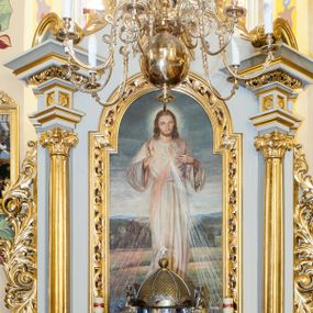 Zdjęcie nr 1: Obraz w kształcie stojącego prostokąta ukazujący stojącego frontalnie Chrystusa. Prawa ręka ułożona w geście błogosławieństwa, lewa wskazuje na promienie odchodzące od piersi. Chrystus ubrany w długą, białą tunikę. Twarz okolona długimi, jasnobrązowymi włosami i krótką, rozwidloną brodą. W tle daleki pagórkowaty krajobraz. 