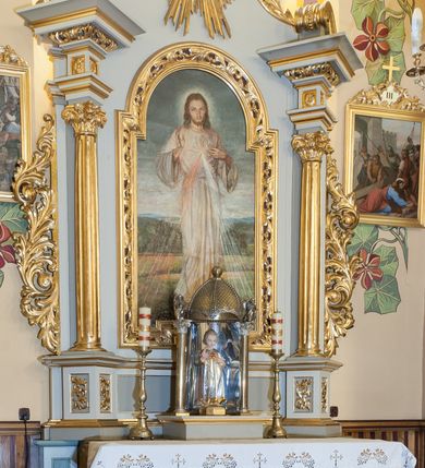 Zdjęcie nr 2: Obraz w kształcie stojącego prostokąta ukazujący stojącego frontalnie Chrystusa. Prawa ręka ułożona w geście błogosławieństwa, lewa wskazuje na promienie odchodzące od piersi. Chrystus ubrany w długą, białą tunikę. Twarz okolona długimi, jasnobrązowymi włosami i krótką, rozwidloną brodą. W tle daleki pagórkowaty krajobraz. 