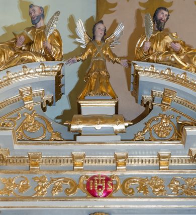 Zdjęcie nr 3: Ołtarz architektoniczny, jednoosiowy, jednokondygnacyjny ze zwieńczeniem. Antepedium z polem prostokątnym o ćwierćkolistych narożach z promianimi wypełnione literami IHS z krzyżem. Na mensie stoi tabernakulum ujęte ukośnie dostawionymi kolumnami i zwieńczone kopułką. Kompozycja nastawy wykorzystuje schemat łuku triumfalnego z arkadą zamknietą półkoliście flankowaną kolumnami stojącymi na postumentach, z kompozytowymi kapitelami dźwigającymi belkowanie. Nad belkowaniem przerwany i wyłamujący się ponad kolumnami, segmentowy przyczółek. W arkadzie obraz z ukrzyżowanym Chrystusem. Na zwieńczeniu dwie siedzące figury ewangelistów (?), brodatych, ubranych w długie suknie i płaszcze, z otwartą księgą i piórem w dłoniach. W partii przerwanego przyczółka stoi na postumencie anioł z rozwartymi rękami, ubrany w długą suknię, z rozpostartymi skrzydłami. 
Cokoły zdobią motywy okuciowe z główkami aniołów. Kolumny w jednej trzeciej ujęte profilowaniem, w dolnej pokryte są okuciowymi taśmami i główkami aniołków, a w górnej wijącą się spiralnie wicią roślinną. Boki nastawy ujmują uszy utworzone z zawijających się taśm. Nad niszą z obrazem główki aniołków. Profile belkowania ozdobione konsolkami i kostkowaniem a fryz wicią roślinną i medalionem z literami IHS. Podobne ornamenty w partii przyczółka. Kolorystyka ołtarza jest szara, kolumny, profile, złocone i srebrzone, twarze aniołków i postaci w zwieńczeniu polichromowane naturalistycznie.  
