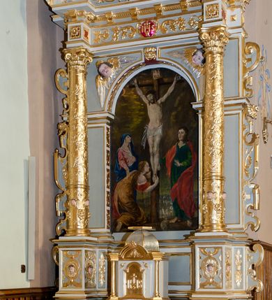 Zdjęcie nr 2: Ołtarz architektoniczny, jednoosiowy, jednokondygnacyjny ze zwieńczeniem. Antepedium z polem prostokątnym o ćwierćkolistych narożach z promianimi wypełnione literami IHS z krzyżem. Na mensie stoi tabernakulum ujęte ukośnie dostawionymi kolumnami i zwieńczone kopułką. Kompozycja nastawy wykorzystuje schemat łuku triumfalnego z arkadą zamknietą półkoliście flankowaną kolumnami stojącymi na postumentach, z kompozytowymi kapitelami dźwigającymi belkowanie. Nad belkowaniem przerwany i wyłamujący się ponad kolumnami, segmentowy przyczółek. W arkadzie obraz z ukrzyżowanym Chrystusem. Na zwieńczeniu dwie siedzące figury ewangelistów (?), brodatych, ubranych w długie suknie i płaszcze, z otwartą księgą i piórem w dłoniach. W partii przerwanego przyczółka stoi na postumencie anioł z rozwartymi rękami, ubrany w długą suknię, z rozpostartymi skrzydłami. 
Cokoły zdobią motywy okuciowe z główkami aniołów. Kolumny w jednej trzeciej ujęte profilowaniem, w dolnej pokryte są okuciowymi taśmami i główkami aniołków, a w górnej wijącą się spiralnie wicią roślinną. Boki nastawy ujmują uszy utworzone z zawijających się taśm. Nad niszą z obrazem główki aniołków. Profile belkowania ozdobione konsolkami i kostkowaniem a fryz wicią roślinną i medalionem z literami IHS. Podobne ornamenty w partii przyczółka. Kolorystyka ołtarza jest szara, kolumny, profile, złocone i srebrzone, twarze aniołków i postaci w zwieńczeniu polichromowane naturalistycznie.  
