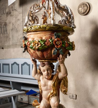 Zdjęcie nr 1: Drewniana, rzeźbiona chrzcielnica ustawiona na okrągłej, ściętej podstawie. Trzon w formie nagiego aniołka stojącego na srebrzonym obłoku, ze złoconymi skrzydłami i draperią przerzucona przez lewe ramię, podtrzymującego oburącz wspartą na głowie czarę w kształcie puklowanej, marmoryzowanej półkuli o wywiniętym kołnierzu. Boki czary ozdobione kwiatową girlandą podwieszoną na wstążkach. Pokrywa ażurowa uformowana z czterech srebrzonych, akantowych wolut wspartych na wieńcu laurowym, w zwieńczeniu promienista gloria z przedstawieniem popiersia Boga Ojca. Pod wolutami, pośrodku pokrywy, rzeźbiona grupa Chrzest Chrystusa. Podstawa i czara marmoryzowana. 