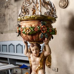 Zdjęcie nr 1: Drewniana, rzeźbiona chrzcielnica ustawiona na okrągłej, ściętej podstawie. Trzon w formie nagiego aniołka stojącego na srebrzonym obłoku, ze złoconymi skrzydłami i draperią przerzucona przez lewe ramię, podtrzymującego oburącz wspartą na głowie czarę w kształcie puklowanej, marmoryzowanej półkuli o wywiniętym kołnierzu. Boki czary ozdobione kwiatową girlandą podwieszoną na wstążkach. Pokrywa ażurowa uformowana z czterech srebrzonych, akantowych wolut wspartych na wieńcu laurowym, w zwieńczeniu promienista gloria z przedstawieniem popiersia Boga Ojca. Pod wolutami, pośrodku pokrywy, rzeźbiona grupa Chrzest Chrystusa. Podstawa i czara marmoryzowana. 