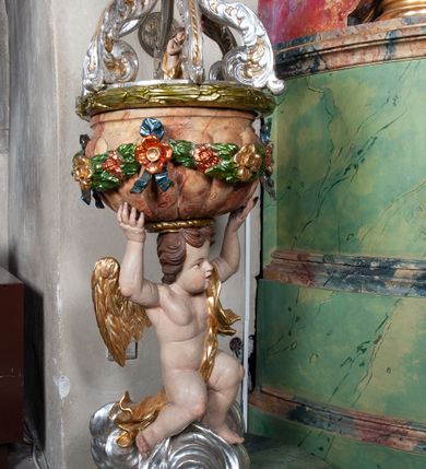 Zdjęcie nr 2: Drewniana, rzeźbiona chrzcielnica ustawiona na okrągłej, ściętej podstawie. Trzon w formie nagiego aniołka stojącego na srebrzonym obłoku, ze złoconymi skrzydłami i draperią przerzucona przez lewe ramię, podtrzymującego oburącz wspartą na głowie czarę w kształcie puklowanej, marmoryzowanej półkuli o wywiniętym kołnierzu. Boki czary ozdobione kwiatową girlandą podwieszoną na wstążkach. Pokrywa ażurowa uformowana z czterech srebrzonych, akantowych wolut wspartych na wieńcu laurowym, w zwieńczeniu promienista gloria z przedstawieniem popiersia Boga Ojca. Pod wolutami, pośrodku pokrywy, rzeźbiona grupa Chrzest Chrystusa. Podstawa i czara marmoryzowana. 
