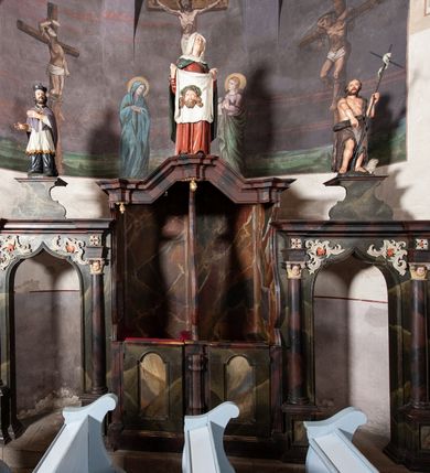 Zdjęcie nr 4: Konfesjonał do słuchania spowiedzi usznej, dwuosobowy, prostopadłościenny, od przodu z parą drzwiczek osadzonych na zawiasach, zwieńczony gzymsem pośrodku wyłamanym trójbocznie. Drzwiczki konfesjonału przedzielone kolumienką, zdobione prostokątnymi płycinami zamkniętymi łukiem półpełnym nadwieszonym. Wewnątrz siedziska oddzielone pionową ścianką o dekoracyjnym wykroju. Płyciny i tylna ściana wnętrza marmoryzowane. Po bokach dwie ścianki parawanowe z prostokątnymi zamkniętymi łukiem kotarowym bramkami dla penitentów. Bramki ujęte pilastrami o kapitelach w formie główki aniołka ze złoconymi skrzydełkami, dźwigającymi uproszczone belkowanie i gzyms występujący nad podporami. W przyłuczach bramek dekoracja malarska. Na gzymsie ustawione rzeźby pełnoplastyczne, polichromowane: w osi środkowej święta Weronika z chustą, w osiach bramek na konsolach o ozdobnym wykroju święty Jan Nepomucen (po lewej) i święty Jan Chrzciciel po prawej. 