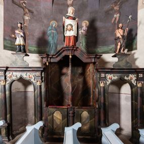 Zdjęcie nr 1: Konfesjonał do słuchania spowiedzi usznej, dwuosobowy, prostopadłościenny, od przodu z parą drzwiczek osadzonych na zawiasach, zwieńczony gzymsem pośrodku wyłamanym trójbocznie. Drzwiczki konfesjonału przedzielone kolumienką, zdobione prostokątnymi płycinami zamkniętymi łukiem półpełnym nadwieszonym. Wewnątrz siedziska oddzielone pionową ścianką o dekoracyjnym wykroju. Płyciny i tylna ściana wnętrza marmoryzowane. Po bokach dwie ścianki parawanowe z prostokątnymi zamkniętymi łukiem kotarowym bramkami dla penitentów. Bramki ujęte pilastrami o kapitelach w formie główki aniołka ze złoconymi skrzydełkami, dźwigającymi uproszczone belkowanie i gzyms występujący nad podporami. W przyłuczach bramek dekoracja malarska. Na gzymsie ustawione rzeźby pełnoplastyczne, polichromowane: w osi środkowej święta Weronika z chustą, w osiach bramek na konsolach o ozdobnym wykroju święty Jan Nepomucen (po lewej) i święty Jan Chrzciciel po prawej. 