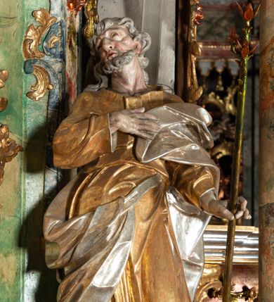 Zdjęcie nr 1: Rzeźba pełnoplastyczna, drążona od tyłu, umieszczona po prawej stronie ołtarza bocznego, przedstawia stojącego na niewysokim, prostopadłościennym, marmoryzowanym postumencie świętego Józefa. Święty ukazany frontalnie, w wyrazistym kontrapoście. Głowa lekko odchylona do tyłu i skręcona w lewo, włosy siwe, falowane, długie, opadające na plecy. Twarz o wyrazistych rysach i krótkim siwym zaroście. Oczy zwrócone w lewo ku górze. Prawa ręka świętego zgięta w łokciu, z dłonią złożoną na piersiach, lewą podtrzymuje kwitnącą różdżkę o długim drzewcu. Święty ubrany w długą, złoconą tunikę i narzucony na nią srebrzony płaszcz, przewieszony przez srebrzony pas opadający na prawe biodro. Fałdy szat układają się równolegle, miejscami spłaszczone, załamujące się. Spod tuniki widoczne stopy w srebrzone sandałach. 