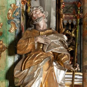 Zdjęcie nr 1: Rzeźba pełnoplastyczna, drążona od tyłu, umieszczona po prawej stronie ołtarza bocznego, przedstawia stojącego na niewysokim, prostopadłościennym, marmoryzowanym postumencie świętego Józefa. Święty ukazany frontalnie, w wyrazistym kontrapoście. Głowa lekko odchylona do tyłu i skręcona w lewo, włosy siwe, falowane, długie, opadające na plecy. Twarz o wyrazistych rysach i krótkim siwym zaroście. Oczy zwrócone w lewo ku górze. Prawa ręka świętego zgięta w łokciu, z dłonią złożoną na piersiach, lewą podtrzymuje kwitnącą różdżkę o długim drzewcu. Święty ubrany w długą, złoconą tunikę i narzucony na nią srebrzony płaszcz, przewieszony przez srebrzony pas opadający na prawe biodro. Fałdy szat układają się równolegle, miejscami spłaszczone, załamujące się. Spod tuniki widoczne stopy w srebrzone sandałach. 