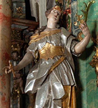 Zdjęcie nr 1: Rzeźba pełnoplastyczna, przyścienna, drążona z tyłu, przedstawiająca stojącą na niewysokim, prostopadłościennym, marmoryzowanym postumencie św. Apolonię. Święta widoczna z profilu, w wyraźnym kontrapoście i lekkim skręcie ciała. Głowa odchylona lekko do tyłu i w lewą stronę. Włosy luźno upięte w kok, z puklem opadającym na prawe ramię. Na głowie złocony diadem zdobiony trzema czerwonymi guzami. Twarz o wyrazistych rysach, wzrok skierowany ku górze. Święta ubrana w spodnią, długą, złoconą i wierzchnią, krótką, srebrzoną tunikę, krótkimi rękawami, ze złoconym gorsetem. Na plecach srebrzony płaszcz, przewieszony przez złocony pas opadający na prawe biodro. Fałdy szat równoległe, spłaszczone, załamujące się. Na stopach srebrzone buty o szpiczastych noskach. W lewej uniesionej ręce gałązka palmowa, w prawej opuszczonej i odwiedzionej w bok obcęgi z zębem. Polichromia partii ciała naturalistyczna. 