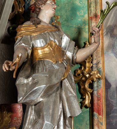 Zdjęcie nr 4: Rzeźba pełnoplastyczna, przyścienna, drążona z tyłu, przedstawiająca stojącą na niewysokim, prostopadłościennym, marmoryzowanym postumencie św. Apolonię. Święta widoczna z profilu, w wyraźnym kontrapoście i lekkim skręcie ciała. Głowa odchylona lekko do tyłu i w lewą stronę. Włosy luźno upięte w kok, z puklem opadającym na prawe ramię. Na głowie złocony diadem zdobiony trzema czerwonymi guzami. Twarz o wyrazistych rysach, wzrok skierowany ku górze. Święta ubrana w spodnią, długą, złoconą i wierzchnią, krótką, srebrzoną tunikę, krótkimi rękawami, ze złoconym gorsetem. Na plecach srebrzony płaszcz, przewieszony przez złocony pas opadający na prawe biodro. Fałdy szat równoległe, spłaszczone, załamujące się. Na stopach srebrzone buty o szpiczastych noskach. W lewej uniesionej ręce gałązka palmowa, w prawej opuszczonej i odwiedzionej w bok obcęgi z zębem. Polichromia partii ciała naturalistyczna. 