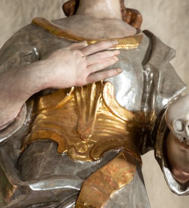 Zdjęcie nr 4: Rzeźba pełnoplastyczna, przyścienna, drążona od tyłu, przedstawiająca stojącą na niewysokim, prostopadłościennym, marmoryzowanym postumencie świętą Rozalię. Święta widoczna z profilu, w wyraźnym kontrapoście, o lekko skręconej sylwetce. Długie, brązowe, falowane włosy luźno spięte na wysokości karku. Na głowie wianek z róż. Twarz o wyrazistych rysach, wzrok skierowany ku górze. Święta ubrana w spodnią, długą, złoconą tunikę i wierzchnią, krótką, srebrzoną, z szerokimi, podwiniętymi rękawami, ze złoconym gorsetem. Na plecach srebrzony płaszcz, podpięty złoconym pasem. Fałdy szat równoległe, spłaszczone, załamujące się. Na stopach srebrzone buty o szpiczastych noskach. W lewej ręce czaszka, prawa uniesiona na wysokość klatki piersiowej. Polichromia partii ciała naturalistyczna.