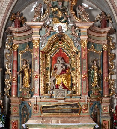 Zdjęcie nr 2: Ołtarz architektoniczny, trójosiowy, z wydzieloną częścią cokołową, jednokondygnacyjny, ze zwieńczeniem. Mensa drewniana tumbowa, ozdobiona nałożonymi płaskorzeźbionymi motywami rocaille’ów i roślinnymi. Po bokach mensy, w części cokołowej dwie bramki obejścia zamknięte półkoliście, w których osadzone na zawiasach drzwi jednoskrzydłowe z płycinami z malowanymi przedstawieniami świętych. W predelli oszklona trumienka z rzeźbą Chrystusa w grobie. Pole środkowe nastawy ujęte dwoma czworobocznymi filarami o kompozytowych kapitelach. Pola boczne wklęsłe i cofnięte od lica pola środkowego flankowane kolumnami o gładkich trzonach i kompozytowych kapitelach, umieszczonych na wysokich dwustopniowych postumentach ustawionych kątowo, zdobionych płycinami. Podpory dźwigają pełne belkowanie i gzyms, przerwane nad polem środkowym. W polu środkowym głęboka nisza w kształcie prostokąta stojącego zamkniętego łukiem wypukło-wklęsłym, w bogatym obramieniu. W niej rzeźba Pieta. Po bokach, na tle filarów, umieszczone dwa okazałe anioły w dynamicznych pozach, z rozłożonymi skrzydłami. Ponad niszą płaskorzeźbiona Chusta św. Weroniki rozpostarta przez dwa uskrzydlone putta ze stopami wspartymi na obłokach. W polach bocznych nastawy sferyczne płyciny w asymetrycznych złoconych obramieniach, na których tle umieszczone na konsolach rzeźby: Św. Jan Ewangelista (po lewej) i Św. Maria Magdalena (po prawej). Zwieńczenie w formie aedikuli ujętej wolutami i zwieńczonej półkoliście. Na tle aedikuli rzeźbione przedstawienie tronującego Boga Ojca, ponad głową którego Arma Christi; wokół putta i uskrzydlone główki aniołków. W osiach podpór rzeźby uskrzydlonych aniołów. Uszaki nastawy rzeźbione, ażurowe, złocone, z motywów rocaille’ów i kwiatów słonecznika. Struktura polichromowana na kolor szaro-niebieski, a trzony filarów i kolumn oraz tła płycin na czerwono, imitując marmur;  detale i ornamenty złocone; szaty rzeźb złocone i srebrzone; karnacje w kolorze naturalnym. 