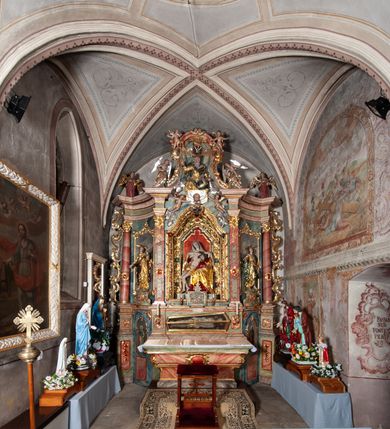 Zdjęcie nr 4: Ołtarz architektoniczny, trójosiowy, z wydzieloną częścią cokołową, jednokondygnacyjny, ze zwieńczeniem. Mensa drewniana tumbowa, ozdobiona nałożonymi płaskorzeźbionymi motywami rocaille’ów i roślinnymi. Po bokach mensy, w części cokołowej dwie bramki obejścia zamknięte półkoliście, w których osadzone na zawiasach drzwi jednoskrzydłowe z płycinami z malowanymi przedstawieniami świętych. W predelli oszklona trumienka z rzeźbą Chrystusa w grobie. Pole środkowe nastawy ujęte dwoma czworobocznymi filarami o kompozytowych kapitelach. Pola boczne wklęsłe i cofnięte od lica pola środkowego flankowane kolumnami o gładkich trzonach i kompozytowych kapitelach, umieszczonych na wysokich dwustopniowych postumentach ustawionych kątowo, zdobionych płycinami. Podpory dźwigają pełne belkowanie i gzyms, przerwane nad polem środkowym. W polu środkowym głęboka nisza w kształcie prostokąta stojącego zamkniętego łukiem wypukło-wklęsłym, w bogatym obramieniu. W niej rzeźba Pieta. Po bokach, na tle filarów, umieszczone dwa okazałe anioły w dynamicznych pozach, z rozłożonymi skrzydłami. Ponad niszą płaskorzeźbiona Chusta św. Weroniki rozpostarta przez dwa uskrzydlone putta ze stopami wspartymi na obłokach. W polach bocznych nastawy sferyczne płyciny w asymetrycznych złoconych obramieniach, na których tle umieszczone na konsolach rzeźby: Św. Jan Ewangelista (po lewej) i Św. Maria Magdalena (po prawej). Zwieńczenie w formie aedikuli ujętej wolutami i zwieńczonej półkoliście. Na tle aedikuli rzeźbione przedstawienie tronującego Boga Ojca, ponad głową którego Arma Christi; wokół putta i uskrzydlone główki aniołków. W osiach podpór rzeźby uskrzydlonych aniołów. Uszaki nastawy rzeźbione, ażurowe, złocone, z motywów rocaille’ów i kwiatów słonecznika. Struktura polichromowana na kolor szaro-niebieski, a trzony filarów i kolumn oraz tła płycin na czerwono, imitując marmur;  detale i ornamenty złocone; szaty rzeźb złocone i srebrzone; karnacje w kolorze naturalnym. 