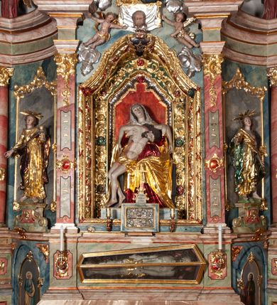 Zdjęcie nr 1: Ołtarz architektoniczny, trójosiowy, z wydzieloną częścią cokołową, jednokondygnacyjny, ze zwieńczeniem. Mensa drewniana tumbowa, ozdobiona nałożonymi płaskorzeźbionymi motywami rocaille’ów i roślinnymi. Po bokach mensy, w części cokołowej dwie bramki obejścia zamknięte półkoliście, w których osadzone na zawiasach drzwi jednoskrzydłowe z płycinami z malowanymi przedstawieniami świętych. W predelli oszklona trumienka z rzeźbą Chrystusa w grobie. Pole środkowe nastawy ujęte dwoma czworobocznymi filarami o kompozytowych kapitelach. Pola boczne wklęsłe i cofnięte od lica pola środkowego flankowane kolumnami o gładkich trzonach i kompozytowych kapitelach, umieszczonych na wysokich dwustopniowych postumentach ustawionych kątowo, zdobionych płycinami. Podpory dźwigają pełne belkowanie i gzyms, przerwane nad polem środkowym. W polu środkowym głęboka nisza w kształcie prostokąta stojącego zamkniętego łukiem wypukło-wklęsłym, w bogatym obramieniu. W niej rzeźba Pieta. Po bokach, na tle filarów, umieszczone dwa okazałe anioły w dynamicznych pozach, z rozłożonymi skrzydłami. Ponad niszą płaskorzeźbiona Chusta św. Weroniki rozpostarta przez dwa uskrzydlone putta ze stopami wspartymi na obłokach. W polach bocznych nastawy sferyczne płyciny w asymetrycznych złoconych obramieniach, na których tle umieszczone na konsolach rzeźby: Św. Jan Ewangelista (po lewej) i Św. Maria Magdalena (po prawej). Zwieńczenie w formie aedikuli ujętej wolutami i zwieńczonej półkoliście. Na tle aedikuli rzeźbione przedstawienie tronującego Boga Ojca, ponad głową którego Arma Christi; wokół putta i uskrzydlone główki aniołków. W osiach podpór rzeźby uskrzydlonych aniołów. Uszaki nastawy rzeźbione, ażurowe, złocone, z motywów rocaille’ów i kwiatów słonecznika. Struktura polichromowana na kolor szaro-niebieski, a trzony filarów i kolumn oraz tła płycin na czerwono, imitując marmur;  detale i ornamenty złocone; szaty rzeźb złocone i srebrzone; karnacje w kolorze naturalnym. 