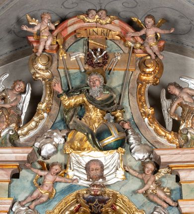 Zdjęcie nr 7: Ołtarz architektoniczny, trójosiowy, z wydzieloną częścią cokołową, jednokondygnacyjny, ze zwieńczeniem. Mensa drewniana tumbowa, ozdobiona nałożonymi płaskorzeźbionymi motywami rocaille’ów i roślinnymi. Po bokach mensy, w części cokołowej dwie bramki obejścia zamknięte półkoliście, w których osadzone na zawiasach drzwi jednoskrzydłowe z płycinami z malowanymi przedstawieniami świętych. W predelli oszklona trumienka z rzeźbą Chrystusa w grobie. Pole środkowe nastawy ujęte dwoma czworobocznymi filarami o kompozytowych kapitelach. Pola boczne wklęsłe i cofnięte od lica pola środkowego flankowane kolumnami o gładkich trzonach i kompozytowych kapitelach, umieszczonych na wysokich dwustopniowych postumentach ustawionych kątowo, zdobionych płycinami. Podpory dźwigają pełne belkowanie i gzyms, przerwane nad polem środkowym. W polu środkowym głęboka nisza w kształcie prostokąta stojącego zamkniętego łukiem wypukło-wklęsłym, w bogatym obramieniu. W niej rzeźba Pieta. Po bokach, na tle filarów, umieszczone dwa okazałe anioły w dynamicznych pozach, z rozłożonymi skrzydłami. Ponad niszą płaskorzeźbiona Chusta św. Weroniki rozpostarta przez dwa uskrzydlone putta ze stopami wspartymi na obłokach. W polach bocznych nastawy sferyczne płyciny w asymetrycznych złoconych obramieniach, na których tle umieszczone na konsolach rzeźby: Św. Jan Ewangelista (po lewej) i Św. Maria Magdalena (po prawej). Zwieńczenie w formie aedikuli ujętej wolutami i zwieńczonej półkoliście. Na tle aedikuli rzeźbione przedstawienie tronującego Boga Ojca, ponad głową którego Arma Christi; wokół putta i uskrzydlone główki aniołków. W osiach podpór rzeźby uskrzydlonych aniołów. Uszaki nastawy rzeźbione, ażurowe, złocone, z motywów rocaille’ów i kwiatów słonecznika. Struktura polichromowana na kolor szaro-niebieski, a trzony filarów i kolumn oraz tła płycin na czerwono, imitując marmur;  detale i ornamenty złocone; szaty rzeźb złocone i srebrzone; karnacje w kolorze naturalnym. 