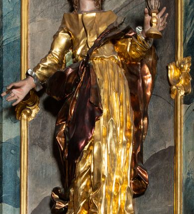 Zdjęcie nr 26: Ołtarz architektoniczny, trójosiowy, z wydzieloną częścią cokołową, jednokondygnacyjny, ze zwieńczeniem. Mensa drewniana tumbowa, ozdobiona nałożonymi płaskorzeźbionymi motywami rocaille’ów i roślinnymi. Po bokach mensy, w części cokołowej dwie bramki obejścia zamknięte półkoliście, w których osadzone na zawiasach drzwi jednoskrzydłowe z płycinami z malowanymi przedstawieniami świętych. W predelli oszklona trumienka z rzeźbą Chrystusa w grobie. Pole środkowe nastawy ujęte dwoma czworobocznymi filarami o kompozytowych kapitelach. Pola boczne wklęsłe i cofnięte od lica pola środkowego flankowane kolumnami o gładkich trzonach i kompozytowych kapitelach, umieszczonych na wysokich dwustopniowych postumentach ustawionych kątowo, zdobionych płycinami. Podpory dźwigają pełne belkowanie i gzyms, przerwane nad polem środkowym. W polu środkowym głęboka nisza w kształcie prostokąta stojącego zamkniętego łukiem wypukło-wklęsłym, w bogatym obramieniu. W niej rzeźba Pieta. Po bokach, na tle filarów, umieszczone dwa okazałe anioły w dynamicznych pozach, z rozłożonymi skrzydłami. Ponad niszą płaskorzeźbiona Chusta św. Weroniki rozpostarta przez dwa uskrzydlone putta ze stopami wspartymi na obłokach. W polach bocznych nastawy sferyczne płyciny w asymetrycznych złoconych obramieniach, na których tle umieszczone na konsolach rzeźby: Św. Jan Ewangelista (po lewej) i Św. Maria Magdalena (po prawej). Zwieńczenie w formie aedikuli ujętej wolutami i zwieńczonej półkoliście. Na tle aedikuli rzeźbione przedstawienie tronującego Boga Ojca, ponad głową którego Arma Christi; wokół putta i uskrzydlone główki aniołków. W osiach podpór rzeźby uskrzydlonych aniołów. Uszaki nastawy rzeźbione, ażurowe, złocone, z motywów rocaille’ów i kwiatów słonecznika. Struktura polichromowana na kolor szaro-niebieski, a trzony filarów i kolumn oraz tła płycin na czerwono, imitując marmur;  detale i ornamenty złocone; szaty rzeźb złocone i srebrzone; karnacje w kolorze naturalnym. 