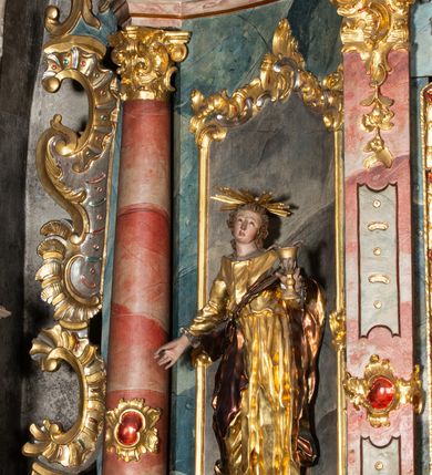 Zdjęcie nr 24: Ołtarz architektoniczny, trójosiowy, z wydzieloną częścią cokołową, jednokondygnacyjny, ze zwieńczeniem. Mensa drewniana tumbowa, ozdobiona nałożonymi płaskorzeźbionymi motywami rocaille’ów i roślinnymi. Po bokach mensy, w części cokołowej dwie bramki obejścia zamknięte półkoliście, w których osadzone na zawiasach drzwi jednoskrzydłowe z płycinami z malowanymi przedstawieniami świętych. W predelli oszklona trumienka z rzeźbą Chrystusa w grobie. Pole środkowe nastawy ujęte dwoma czworobocznymi filarami o kompozytowych kapitelach. Pola boczne wklęsłe i cofnięte od lica pola środkowego flankowane kolumnami o gładkich trzonach i kompozytowych kapitelach, umieszczonych na wysokich dwustopniowych postumentach ustawionych kątowo, zdobionych płycinami. Podpory dźwigają pełne belkowanie i gzyms, przerwane nad polem środkowym. W polu środkowym głęboka nisza w kształcie prostokąta stojącego zamkniętego łukiem wypukło-wklęsłym, w bogatym obramieniu. W niej rzeźba Pieta. Po bokach, na tle filarów, umieszczone dwa okazałe anioły w dynamicznych pozach, z rozłożonymi skrzydłami. Ponad niszą płaskorzeźbiona Chusta św. Weroniki rozpostarta przez dwa uskrzydlone putta ze stopami wspartymi na obłokach. W polach bocznych nastawy sferyczne płyciny w asymetrycznych złoconych obramieniach, na których tle umieszczone na konsolach rzeźby: Św. Jan Ewangelista (po lewej) i Św. Maria Magdalena (po prawej). Zwieńczenie w formie aedikuli ujętej wolutami i zwieńczonej półkoliście. Na tle aedikuli rzeźbione przedstawienie tronującego Boga Ojca, ponad głową którego Arma Christi; wokół putta i uskrzydlone główki aniołków. W osiach podpór rzeźby uskrzydlonych aniołów. Uszaki nastawy rzeźbione, ażurowe, złocone, z motywów rocaille’ów i kwiatów słonecznika. Struktura polichromowana na kolor szaro-niebieski, a trzony filarów i kolumn oraz tła płycin na czerwono, imitując marmur;  detale i ornamenty złocone; szaty rzeźb złocone i srebrzone; karnacje w kolorze naturalnym. 