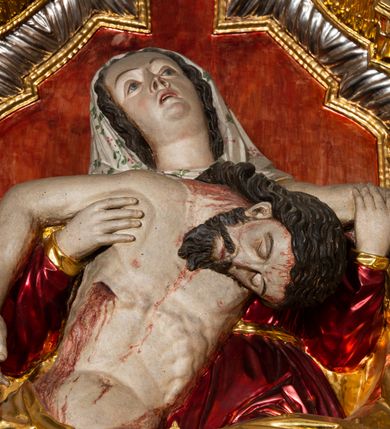 Zdjęcie nr 17: Ołtarz architektoniczny, trójosiowy, z wydzieloną częścią cokołową, jednokondygnacyjny, ze zwieńczeniem. Mensa drewniana tumbowa, ozdobiona nałożonymi płaskorzeźbionymi motywami rocaille’ów i roślinnymi. Po bokach mensy, w części cokołowej dwie bramki obejścia zamknięte półkoliście, w których osadzone na zawiasach drzwi jednoskrzydłowe z płycinami z malowanymi przedstawieniami świętych. W predelli oszklona trumienka z rzeźbą Chrystusa w grobie. Pole środkowe nastawy ujęte dwoma czworobocznymi filarami o kompozytowych kapitelach. Pola boczne wklęsłe i cofnięte od lica pola środkowego flankowane kolumnami o gładkich trzonach i kompozytowych kapitelach, umieszczonych na wysokich dwustopniowych postumentach ustawionych kątowo, zdobionych płycinami. Podpory dźwigają pełne belkowanie i gzyms, przerwane nad polem środkowym. W polu środkowym głęboka nisza w kształcie prostokąta stojącego zamkniętego łukiem wypukło-wklęsłym, w bogatym obramieniu. W niej rzeźba Pieta. Po bokach, na tle filarów, umieszczone dwa okazałe anioły w dynamicznych pozach, z rozłożonymi skrzydłami. Ponad niszą płaskorzeźbiona Chusta św. Weroniki rozpostarta przez dwa uskrzydlone putta ze stopami wspartymi na obłokach. W polach bocznych nastawy sferyczne płyciny w asymetrycznych złoconych obramieniach, na których tle umieszczone na konsolach rzeźby: Św. Jan Ewangelista (po lewej) i Św. Maria Magdalena (po prawej). Zwieńczenie w formie aedikuli ujętej wolutami i zwieńczonej półkoliście. Na tle aedikuli rzeźbione przedstawienie tronującego Boga Ojca, ponad głową którego Arma Christi; wokół putta i uskrzydlone główki aniołków. W osiach podpór rzeźby uskrzydlonych aniołów. Uszaki nastawy rzeźbione, ażurowe, złocone, z motywów rocaille’ów i kwiatów słonecznika. Struktura polichromowana na kolor szaro-niebieski, a trzony filarów i kolumn oraz tła płycin na czerwono, imitując marmur;  detale i ornamenty złocone; szaty rzeźb złocone i srebrzone; karnacje w kolorze naturalnym. 