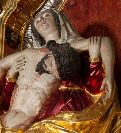 Zdjęcie nr 16: Ołtarz architektoniczny, trójosiowy, z wydzieloną częścią cokołową, jednokondygnacyjny, ze zwieńczeniem. Mensa drewniana tumbowa, ozdobiona nałożonymi płaskorzeźbionymi motywami rocaille’ów i roślinnymi. Po bokach mensy, w części cokołowej dwie bramki obejścia zamknięte półkoliście, w których osadzone na zawiasach drzwi jednoskrzydłowe z płycinami z malowanymi przedstawieniami świętych. W predelli oszklona trumienka z rzeźbą Chrystusa w grobie. Pole środkowe nastawy ujęte dwoma czworobocznymi filarami o kompozytowych kapitelach. Pola boczne wklęsłe i cofnięte od lica pola środkowego flankowane kolumnami o gładkich trzonach i kompozytowych kapitelach, umieszczonych na wysokich dwustopniowych postumentach ustawionych kątowo, zdobionych płycinami. Podpory dźwigają pełne belkowanie i gzyms, przerwane nad polem środkowym. W polu środkowym głęboka nisza w kształcie prostokąta stojącego zamkniętego łukiem wypukło-wklęsłym, w bogatym obramieniu. W niej rzeźba Pieta. Po bokach, na tle filarów, umieszczone dwa okazałe anioły w dynamicznych pozach, z rozłożonymi skrzydłami. Ponad niszą płaskorzeźbiona Chusta św. Weroniki rozpostarta przez dwa uskrzydlone putta ze stopami wspartymi na obłokach. W polach bocznych nastawy sferyczne płyciny w asymetrycznych złoconych obramieniach, na których tle umieszczone na konsolach rzeźby: Św. Jan Ewangelista (po lewej) i Św. Maria Magdalena (po prawej). Zwieńczenie w formie aedikuli ujętej wolutami i zwieńczonej półkoliście. Na tle aedikuli rzeźbione przedstawienie tronującego Boga Ojca, ponad głową którego Arma Christi; wokół putta i uskrzydlone główki aniołków. W osiach podpór rzeźby uskrzydlonych aniołów. Uszaki nastawy rzeźbione, ażurowe, złocone, z motywów rocaille’ów i kwiatów słonecznika. Struktura polichromowana na kolor szaro-niebieski, a trzony filarów i kolumn oraz tła płycin na czerwono, imitując marmur;  detale i ornamenty złocone; szaty rzeźb złocone i srebrzone; karnacje w kolorze naturalnym. 