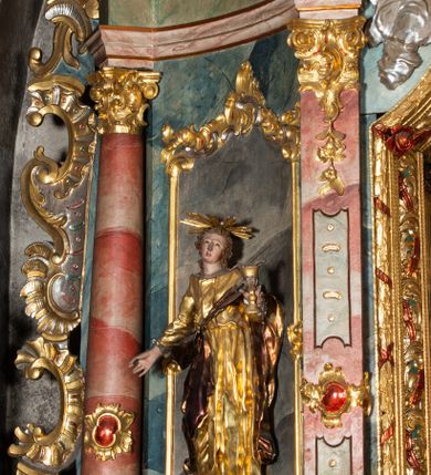 Zdjęcie nr 25: Ołtarz architektoniczny, trójosiowy, z wydzieloną częścią cokołową, jednokondygnacyjny, ze zwieńczeniem. Mensa drewniana tumbowa, ozdobiona nałożonymi płaskorzeźbionymi motywami rocaille’ów i roślinnymi. Po bokach mensy, w części cokołowej dwie bramki obejścia zamknięte półkoliście, w których osadzone na zawiasach drzwi jednoskrzydłowe z płycinami z malowanymi przedstawieniami świętych. W predelli oszklona trumienka z rzeźbą Chrystusa w grobie. Pole środkowe nastawy ujęte dwoma czworobocznymi filarami o kompozytowych kapitelach. Pola boczne wklęsłe i cofnięte od lica pola środkowego flankowane kolumnami o gładkich trzonach i kompozytowych kapitelach, umieszczonych na wysokich dwustopniowych postumentach ustawionych kątowo, zdobionych płycinami. Podpory dźwigają pełne belkowanie i gzyms, przerwane nad polem środkowym. W polu środkowym głęboka nisza w kształcie prostokąta stojącego zamkniętego łukiem wypukło-wklęsłym, w bogatym obramieniu. W niej rzeźba Pieta. Po bokach, na tle filarów, umieszczone dwa okazałe anioły w dynamicznych pozach, z rozłożonymi skrzydłami. Ponad niszą płaskorzeźbiona Chusta św. Weroniki rozpostarta przez dwa uskrzydlone putta ze stopami wspartymi na obłokach. W polach bocznych nastawy sferyczne płyciny w asymetrycznych złoconych obramieniach, na których tle umieszczone na konsolach rzeźby: Św. Jan Ewangelista (po lewej) i Św. Maria Magdalena (po prawej). Zwieńczenie w formie aedikuli ujętej wolutami i zwieńczonej półkoliście. Na tle aedikuli rzeźbione przedstawienie tronującego Boga Ojca, ponad głową którego Arma Christi; wokół putta i uskrzydlone główki aniołków. W osiach podpór rzeźby uskrzydlonych aniołów. Uszaki nastawy rzeźbione, ażurowe, złocone, z motywów rocaille’ów i kwiatów słonecznika. Struktura polichromowana na kolor szaro-niebieski, a trzony filarów i kolumn oraz tła płycin na czerwono, imitując marmur;  detale i ornamenty złocone; szaty rzeźb złocone i srebrzone; karnacje w kolorze naturalnym. 