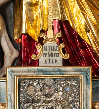 Zdjęcie nr 19: Ołtarz architektoniczny, trójosiowy, z wydzieloną częścią cokołową, jednokondygnacyjny, ze zwieńczeniem. Mensa drewniana tumbowa, ozdobiona nałożonymi płaskorzeźbionymi motywami rocaille’ów i roślinnymi. Po bokach mensy, w części cokołowej dwie bramki obejścia zamknięte półkoliście, w których osadzone na zawiasach drzwi jednoskrzydłowe z płycinami z malowanymi przedstawieniami świętych. W predelli oszklona trumienka z rzeźbą Chrystusa w grobie. Pole środkowe nastawy ujęte dwoma czworobocznymi filarami o kompozytowych kapitelach. Pola boczne wklęsłe i cofnięte od lica pola środkowego flankowane kolumnami o gładkich trzonach i kompozytowych kapitelach, umieszczonych na wysokich dwustopniowych postumentach ustawionych kątowo, zdobionych płycinami. Podpory dźwigają pełne belkowanie i gzyms, przerwane nad polem środkowym. W polu środkowym głęboka nisza w kształcie prostokąta stojącego zamkniętego łukiem wypukło-wklęsłym, w bogatym obramieniu. W niej rzeźba Pieta. Po bokach, na tle filarów, umieszczone dwa okazałe anioły w dynamicznych pozach, z rozłożonymi skrzydłami. Ponad niszą płaskorzeźbiona Chusta św. Weroniki rozpostarta przez dwa uskrzydlone putta ze stopami wspartymi na obłokach. W polach bocznych nastawy sferyczne płyciny w asymetrycznych złoconych obramieniach, na których tle umieszczone na konsolach rzeźby: Św. Jan Ewangelista (po lewej) i Św. Maria Magdalena (po prawej). Zwieńczenie w formie aedikuli ujętej wolutami i zwieńczonej półkoliście. Na tle aedikuli rzeźbione przedstawienie tronującego Boga Ojca, ponad głową którego Arma Christi; wokół putta i uskrzydlone główki aniołków. W osiach podpór rzeźby uskrzydlonych aniołów. Uszaki nastawy rzeźbione, ażurowe, złocone, z motywów rocaille’ów i kwiatów słonecznika. Struktura polichromowana na kolor szaro-niebieski, a trzony filarów i kolumn oraz tła płycin na czerwono, imitując marmur;  detale i ornamenty złocone; szaty rzeźb złocone i srebrzone; karnacje w kolorze naturalnym. 