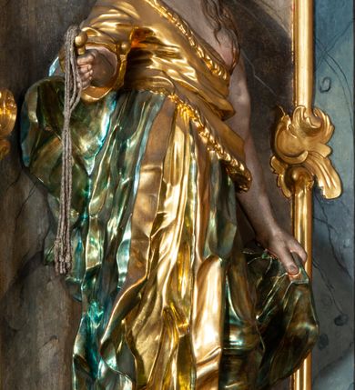 Zdjęcie nr 29: Ołtarz architektoniczny, trójosiowy, z wydzieloną częścią cokołową, jednokondygnacyjny, ze zwieńczeniem. Mensa drewniana tumbowa, ozdobiona nałożonymi płaskorzeźbionymi motywami rocaille’ów i roślinnymi. Po bokach mensy, w części cokołowej dwie bramki obejścia zamknięte półkoliście, w których osadzone na zawiasach drzwi jednoskrzydłowe z płycinami z malowanymi przedstawieniami świętych. W predelli oszklona trumienka z rzeźbą Chrystusa w grobie. Pole środkowe nastawy ujęte dwoma czworobocznymi filarami o kompozytowych kapitelach. Pola boczne wklęsłe i cofnięte od lica pola środkowego flankowane kolumnami o gładkich trzonach i kompozytowych kapitelach, umieszczonych na wysokich dwustopniowych postumentach ustawionych kątowo, zdobionych płycinami. Podpory dźwigają pełne belkowanie i gzyms, przerwane nad polem środkowym. W polu środkowym głęboka nisza w kształcie prostokąta stojącego zamkniętego łukiem wypukło-wklęsłym, w bogatym obramieniu. W niej rzeźba Pieta. Po bokach, na tle filarów, umieszczone dwa okazałe anioły w dynamicznych pozach, z rozłożonymi skrzydłami. Ponad niszą płaskorzeźbiona Chusta św. Weroniki rozpostarta przez dwa uskrzydlone putta ze stopami wspartymi na obłokach. W polach bocznych nastawy sferyczne płyciny w asymetrycznych złoconych obramieniach, na których tle umieszczone na konsolach rzeźby: Św. Jan Ewangelista (po lewej) i Św. Maria Magdalena (po prawej). Zwieńczenie w formie aedikuli ujętej wolutami i zwieńczonej półkoliście. Na tle aedikuli rzeźbione przedstawienie tronującego Boga Ojca, ponad głową którego Arma Christi; wokół putta i uskrzydlone główki aniołków. W osiach podpór rzeźby uskrzydlonych aniołów. Uszaki nastawy rzeźbione, ażurowe, złocone, z motywów rocaille’ów i kwiatów słonecznika. Struktura polichromowana na kolor szaro-niebieski, a trzony filarów i kolumn oraz tła płycin na czerwono, imitując marmur;  detale i ornamenty złocone; szaty rzeźb złocone i srebrzone; karnacje w kolorze naturalnym. 