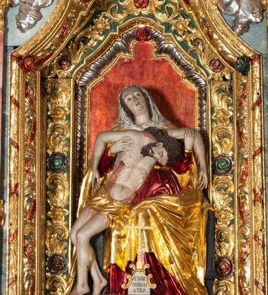 Zdjęcie nr 15: Ołtarz architektoniczny, trójosiowy, z wydzieloną częścią cokołową, jednokondygnacyjny, ze zwieńczeniem. Mensa drewniana tumbowa, ozdobiona nałożonymi płaskorzeźbionymi motywami rocaille’ów i roślinnymi. Po bokach mensy, w części cokołowej dwie bramki obejścia zamknięte półkoliście, w których osadzone na zawiasach drzwi jednoskrzydłowe z płycinami z malowanymi przedstawieniami świętych. W predelli oszklona trumienka z rzeźbą Chrystusa w grobie. Pole środkowe nastawy ujęte dwoma czworobocznymi filarami o kompozytowych kapitelach. Pola boczne wklęsłe i cofnięte od lica pola środkowego flankowane kolumnami o gładkich trzonach i kompozytowych kapitelach, umieszczonych na wysokich dwustopniowych postumentach ustawionych kątowo, zdobionych płycinami. Podpory dźwigają pełne belkowanie i gzyms, przerwane nad polem środkowym. W polu środkowym głęboka nisza w kształcie prostokąta stojącego zamkniętego łukiem wypukło-wklęsłym, w bogatym obramieniu. W niej rzeźba Pieta. Po bokach, na tle filarów, umieszczone dwa okazałe anioły w dynamicznych pozach, z rozłożonymi skrzydłami. Ponad niszą płaskorzeźbiona Chusta św. Weroniki rozpostarta przez dwa uskrzydlone putta ze stopami wspartymi na obłokach. W polach bocznych nastawy sferyczne płyciny w asymetrycznych złoconych obramieniach, na których tle umieszczone na konsolach rzeźby: Św. Jan Ewangelista (po lewej) i Św. Maria Magdalena (po prawej). Zwieńczenie w formie aedikuli ujętej wolutami i zwieńczonej półkoliście. Na tle aedikuli rzeźbione przedstawienie tronującego Boga Ojca, ponad głową którego Arma Christi; wokół putta i uskrzydlone główki aniołków. W osiach podpór rzeźby uskrzydlonych aniołów. Uszaki nastawy rzeźbione, ażurowe, złocone, z motywów rocaille’ów i kwiatów słonecznika. Struktura polichromowana na kolor szaro-niebieski, a trzony filarów i kolumn oraz tła płycin na czerwono, imitując marmur;  detale i ornamenty złocone; szaty rzeźb złocone i srebrzone; karnacje w kolorze naturalnym. 