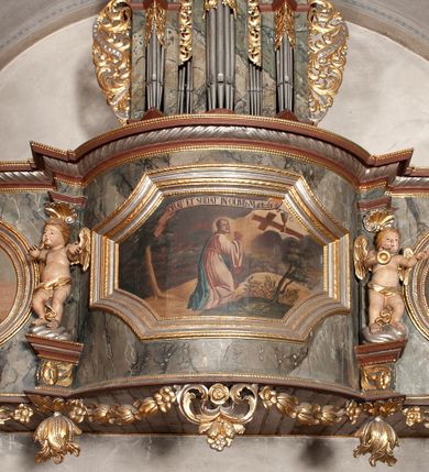 Zdjęcie nr 1: Prospekt organowy jednokondygnacyjny, trójosiowy, złożony z trzech prostopadłościennych segmentów z ryzalitami; środkowy najwyższy, na rzucie półkolisty, skrajne niższe, dwuboczne, dźwigające odcinki belkowania i profilowanego gzymsu; w zwieńczeniu owalne medaliony ujęte liśćmi akantu. Ryzality wypełnione piszczałkami, częściowo przesłoniętymi ażurową dekoracją. Prospekt ujęty ażurowymi uszakami z motywów roślinnych. Struktura polichromowana w kolorze drewna, ornamenty złocone.
