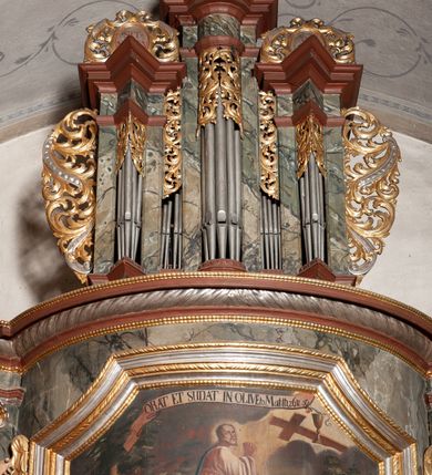Zdjęcie nr 2: Prospekt organowy jednokondygnacyjny, trójosiowy, złożony z trzech prostopadłościennych segmentów z ryzalitami; środkowy najwyższy, na rzucie półkolisty, skrajne niższe, dwuboczne, dźwigające odcinki belkowania i profilowanego gzymsu; w zwieńczeniu owalne medaliony ujęte liśćmi akantu. Ryzality wypełnione piszczałkami, częściowo przesłoniętymi ażurową dekoracją. Prospekt ujęty ażurowymi uszakami z motywów roślinnych. Struktura polichromowana w kolorze drewna, ornamenty złocone.
