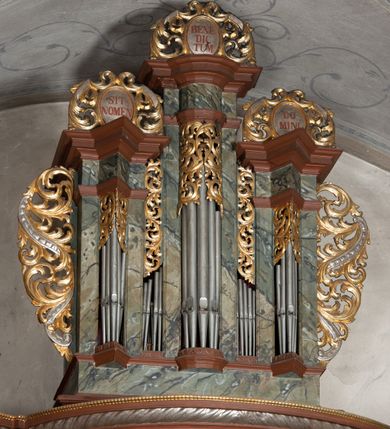 Zdjęcie nr 3: Prospekt organowy jednokondygnacyjny, trójosiowy, złożony z trzech prostopadłościennych segmentów z ryzalitami; środkowy najwyższy, na rzucie półkolisty, skrajne niższe, dwuboczne, dźwigające odcinki belkowania i profilowanego gzymsu; w zwieńczeniu owalne medaliony ujęte liśćmi akantu. Ryzality wypełnione piszczałkami, częściowo przesłoniętymi ażurową dekoracją. Prospekt ujęty ażurowymi uszakami z motywów roślinnych. Struktura polichromowana w kolorze drewna, ornamenty złocone.
