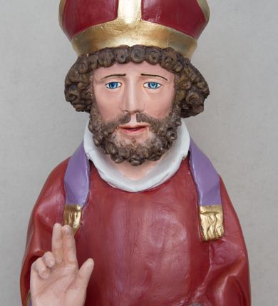 Zdjęcie nr 2: Rzeźba św. Mikołaja ustawiona na prostopadłościennej podstawie feretronu, zaopatrzonej w otwory na drążki do noszenia. Święty ukazany jest w postawie stojącej na niskim cokole, frontalnie, w statycznej pozie z rękami wyciągniętymi do przodu, ściśle przylegającymi do sylwetki: prawą wykonuje gest błogosławieństwa, a w lewej trzyma księgę i trzy srebrzone kule. Ma szeroką twarz z długim i prostym nosem, wzrokiem skierowanym na wprost oraz bujną brodę skręconą w bujne, niewielkie loki. Włosy krótkie, kędzierzawe, jasnobrązowe, ułożone w drobne loki. Święty ubrany jest w fioletową albę, czerwony ornat  z fioletową podszewką i złotym obrzeżem, na głowie ma czerwoną infułę, a na szyi biały humerał i krótką, fioletową stułę, na stopach czarne buty. Draperia szat jest silnie łamana w dolnej partii o ostrych krawędziach. Polichromia w odsłoniętych partiach ciała naturalistyczna, podstawa feretronu marmoryzowana na kolor jasnozielony, detale złocone i srebrzone.

