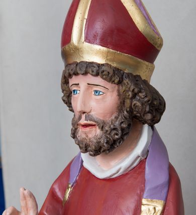 Zdjęcie nr 4: Rzeźba św. Mikołaja ustawiona na prostopadłościennej podstawie feretronu, zaopatrzonej w otwory na drążki do noszenia. Święty ukazany jest w postawie stojącej na niskim cokole, frontalnie, w statycznej pozie z rękami wyciągniętymi do przodu, ściśle przylegającymi do sylwetki: prawą wykonuje gest błogosławieństwa, a w lewej trzyma księgę i trzy srebrzone kule. Ma szeroką twarz z długim i prostym nosem, wzrokiem skierowanym na wprost oraz bujną brodę skręconą w bujne, niewielkie loki. Włosy krótkie, kędzierzawe, jasnobrązowe, ułożone w drobne loki. Święty ubrany jest w fioletową albę, czerwony ornat  z fioletową podszewką i złotym obrzeżem, na głowie ma czerwoną infułę, a na szyi biały humerał i krótką, fioletową stułę, na stopach czarne buty. Draperia szat jest silnie łamana w dolnej partii o ostrych krawędziach. Polichromia w odsłoniętych partiach ciała naturalistyczna, podstawa feretronu marmoryzowana na kolor jasnozielony, detale złocone i srebrzone.

