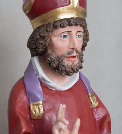 Zdjęcie nr 3: Rzeźba św. Mikołaja ustawiona na prostopadłościennej podstawie feretronu, zaopatrzonej w otwory na drążki do noszenia. Święty ukazany jest w postawie stojącej na niskim cokole, frontalnie, w statycznej pozie z rękami wyciągniętymi do przodu, ściśle przylegającymi do sylwetki: prawą wykonuje gest błogosławieństwa, a w lewej trzyma księgę i trzy srebrzone kule. Ma szeroką twarz z długim i prostym nosem, wzrokiem skierowanym na wprost oraz bujną brodę skręconą w bujne, niewielkie loki. Włosy krótkie, kędzierzawe, jasnobrązowe, ułożone w drobne loki. Święty ubrany jest w fioletową albę, czerwony ornat  z fioletową podszewką i złotym obrzeżem, na głowie ma czerwoną infułę, a na szyi biały humerał i krótką, fioletową stułę, na stopach czarne buty. Draperia szat jest silnie łamana w dolnej partii o ostrych krawędziach. Polichromia w odsłoniętych partiach ciała naturalistyczna, podstawa feretronu marmoryzowana na kolor jasnozielony, detale złocone i srebrzone.

