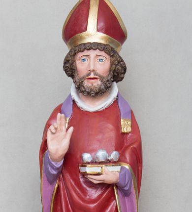 Zdjęcie nr 1: Rzeźba św. Mikołaja ustawiona na prostopadłościennej podstawie feretronu, zaopatrzonej w otwory na drążki do noszenia. Święty ukazany jest w postawie stojącej na niskim cokole, frontalnie, w statycznej pozie z rękami wyciągniętymi do przodu, ściśle przylegającymi do sylwetki: prawą wykonuje gest błogosławieństwa, a w lewej trzyma księgę i trzy srebrzone kule. Ma szeroką twarz z długim i prostym nosem, wzrokiem skierowanym na wprost oraz bujną brodę skręconą w bujne, niewielkie loki. Włosy krótkie, kędzierzawe, jasnobrązowe, ułożone w drobne loki. Święty ubrany jest w fioletową albę, czerwony ornat  z fioletową podszewką i złotym obrzeżem, na głowie ma czerwoną infułę, a na szyi biały humerał i krótką, fioletową stułę, na stopach czarne buty. Draperia szat jest silnie łamana w dolnej partii o ostrych krawędziach. Polichromia w odsłoniętych partiach ciała naturalistyczna, podstawa feretronu marmoryzowana na kolor jasnozielony, detale złocone i srebrzone.

