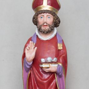 Zdjęcie nr 1: Rzeźba św. Mikołaja ustawiona na prostopadłościennej podstawie feretronu, zaopatrzonej w otwory na drążki do noszenia. Święty ukazany jest w postawie stojącej na niskim cokole, frontalnie, w statycznej pozie z rękami wyciągniętymi do przodu, ściśle przylegającymi do sylwetki: prawą wykonuje gest błogosławieństwa, a w lewej trzyma księgę i trzy srebrzone kule. Ma szeroką twarz z długim i prostym nosem, wzrokiem skierowanym na wprost oraz bujną brodę skręconą w bujne, niewielkie loki. Włosy krótkie, kędzierzawe, jasnobrązowe, ułożone w drobne loki. Święty ubrany jest w fioletową albę, czerwony ornat  z fioletową podszewką i złotym obrzeżem, na głowie ma czerwoną infułę, a na szyi biały humerał i krótką, fioletową stułę, na stopach czarne buty. Draperia szat jest silnie łamana w dolnej partii o ostrych krawędziach. Polichromia w odsłoniętych partiach ciała naturalistyczna, podstawa feretronu marmoryzowana na kolor jasnozielony, detale złocone i srebrzone.

