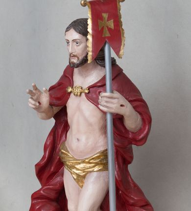 Zdjęcie nr 2: Rzeźba Chrystusa Zmartwychwstałego wolnostojąca, ustawiona na niskim, czarnym cokole o ściętych narożach. Jezus ukazany w całej postaci ustawiony frontalnie na obłoku, w delikatnym kontrapoście z lewą nogą ugiętą w kolanie, prawą ręką uniesioną na wysokość piersi czyni gest błogosławieństwa, w lewej trzyma chorągiew o długim drzewcu, zakończonym złoconym krzyżem oraz z zawieszoną czerwoną tkaniną ze złoconym krzyżem w polu. Chrystus ma podłużną twarz z długim i wąskim nosem, wzrokiem skierowanym w dal, okoloną krótką i ciemną brodą i długimi oraz falowanymi włosami, które opadają na plecy i ramiona. Jest ubrany w złocone perizonium, czerwony płaszcz, spięty pod szyją złotą klamrą, odsłaniający nagi tors, otaczający figurę, silnie drapowany. Na dłoniach, bosych stopach i boku zaznaczone są ślady męki. Polichromia ciała jest naturalistyczna.