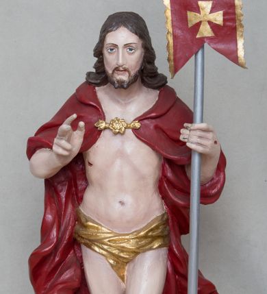 Zdjęcie nr 1: Rzeźba Chrystusa Zmartwychwstałego wolnostojąca, ustawiona na niskim, czarnym cokole o ściętych narożach. Jezus ukazany w całej postaci ustawiony frontalnie na obłoku, w delikatnym kontrapoście z lewą nogą ugiętą w kolanie, prawą ręką uniesioną na wysokość piersi czyni gest błogosławieństwa, w lewej trzyma chorągiew o długim drzewcu, zakończonym złoconym krzyżem oraz z zawieszoną czerwoną tkaniną ze złoconym krzyżem w polu. Chrystus ma podłużną twarz z długim i wąskim nosem, wzrokiem skierowanym w dal, okoloną krótką i ciemną brodą i długimi oraz falowanymi włosami, które opadają na plecy i ramiona. Jest ubrany w złocone perizonium, czerwony płaszcz, spięty pod szyją złotą klamrą, odsłaniający nagi tors, otaczający figurę, silnie drapowany. Na dłoniach, bosych stopach i boku zaznaczone są ślady męki. Polichromia ciała jest naturalistyczna.