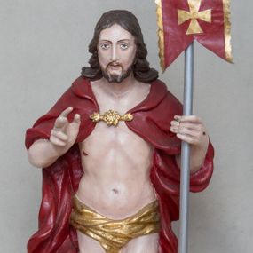 Zdjęcie nr 1: Rzeźba Chrystusa Zmartwychwstałego wolnostojąca, ustawiona na niskim, czarnym cokole o ściętych narożach. Jezus ukazany w całej postaci ustawiony frontalnie na obłoku, w delikatnym kontrapoście z lewą nogą ugiętą w kolanie, prawą ręką uniesioną na wysokość piersi czyni gest błogosławieństwa, w lewej trzyma chorągiew o długim drzewcu, zakończonym złoconym krzyżem oraz z zawieszoną czerwoną tkaniną ze złoconym krzyżem w polu. Chrystus ma podłużną twarz z długim i wąskim nosem, wzrokiem skierowanym w dal, okoloną krótką i ciemną brodą i długimi oraz falowanymi włosami, które opadają na plecy i ramiona. Jest ubrany w złocone perizonium, czerwony płaszcz, spięty pod szyją złotą klamrą, odsłaniający nagi tors, otaczający figurę, silnie drapowany. Na dłoniach, bosych stopach i boku zaznaczone są ślady męki. Polichromia ciała jest naturalistyczna.