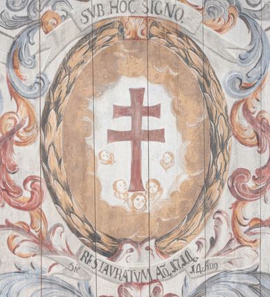 Zdjęcie nr 17: Polichromia umieszczona na stropie nawy głównej. W polu środkowym scena Wniebowzięcia Najświętszej Marii Panny umieszczona w kwadratowej ramie o bokach zamkniętych łukami nadwieszonymi. Rama profilowana, zdobiona rzędem pojedynczych liści. Matka Boska unosząca się na obłokach z szeroko rozłożonymi na boki rękami, prawą wzniesioną do góry. Twarz owalna z długim nosem, silnie zaciśniętymi ustami i dużymi oczami, skierowanymi ku górze. Włosy jasne, kręcone, schowane pod żółtym welonem, którego jeden koniec rozwiany jest wzdłuż prawej ręki. Matka Boska ubrana jest w szeroką, różową suknię, przewiązaną w talii pomarańczową tasiemką oraz błękitny płaszcz, założony za lewe ramię i rozwiany z tyłu. Nad głową Matki Boskiej aniołki unoszą insygnia królewskie: koronę, berło, a także gałązkę białej lilii. Poniżej grupy aniołów podtrzymujące obłoki. Dokoła ramy bujne sploty różnokolorowego akantu przeplecionego karbowaną wstęgą, owijające również ramę ze sceną Wniebowzięcia. W dolnej części ukazane są ponadto dwa anioły podtrzymujące białą wstęgę z napisem: „ASSVMPTA EST MARIA IN COELVM, GAV, / DENT ANGELI, LAVDANTES BENEDICVNT / DOMINVM.” Z kolei z drugiej strony stropu umieszczony jest owalny medalion, obwiedziony liśćmi wawrzynu, w polu którego umieszczone jest czerwone godło zakonu Bożogrobowców oraz małe uskrzydlone główki aniołków. Powyżej na białej wstędze umieszczony jest napis: „SVB HOC SIGNO.”, a poniżej: „RESTAVRATVM A(NN)O (DOMINI) 1714 / Die 14 Aug(st)”. Całość stropu obwiedziona jest dookoła pasem przeważnie prostokątnych kasetonów, wypełnionych różnokolorowymi liśćmi akantu, a także od zewnątrz szerokim pasem dekoracyjnym z brązowych liści wawrzynu. Pasy na bokach wyłamane półkolistymi wypustkami, w których umieszczone są duże muszle palmetowe. Kolorystyka stonowana, jasna z przewagą błękitu i czerwieni.