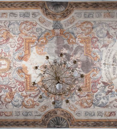 Zdjęcie nr 15: Polichromia umieszczona na stropie nawy głównej. W polu środkowym scena Wniebowzięcia Najświętszej Marii Panny umieszczona w kwadratowej ramie o bokach zamkniętych łukami nadwieszonymi. Rama profilowana, zdobiona rzędem pojedynczych liści. Matka Boska unosząca się na obłokach z szeroko rozłożonymi na boki rękami, prawą wzniesioną do góry. Twarz owalna z długim nosem, silnie zaciśniętymi ustami i dużymi oczami, skierowanymi ku górze. Włosy jasne, kręcone, schowane pod żółtym welonem, którego jeden koniec rozwiany jest wzdłuż prawej ręki. Matka Boska ubrana jest w szeroką, różową suknię, przewiązaną w talii pomarańczową tasiemką oraz błękitny płaszcz, założony za lewe ramię i rozwiany z tyłu. Nad głową Matki Boskiej aniołki unoszą insygnia królewskie: koronę, berło, a także gałązkę białej lilii. Poniżej grupy aniołów podtrzymujące obłoki. Dokoła ramy bujne sploty różnokolorowego akantu przeplecionego karbowaną wstęgą, owijające również ramę ze sceną Wniebowzięcia. W dolnej części ukazane są ponadto dwa anioły podtrzymujące białą wstęgę z napisem: „ASSVMPTA EST MARIA IN COELVM, GAV, / DENT ANGELI, LAVDANTES BENEDICVNT / DOMINVM.” Z kolei z drugiej strony stropu umieszczony jest owalny medalion, obwiedziony liśćmi wawrzynu, w polu którego umieszczone jest czerwone godło zakonu Bożogrobowców oraz małe uskrzydlone główki aniołków. Powyżej na białej wstędze umieszczony jest napis: „SVB HOC SIGNO.”, a poniżej: „RESTAVRATVM A(NN)O (DOMINI) 1714 / Die 14 Aug(st)”. Całość stropu obwiedziona jest dookoła pasem przeważnie prostokątnych kasetonów, wypełnionych różnokolorowymi liśćmi akantu, a także od zewnątrz szerokim pasem dekoracyjnym z brązowych liści wawrzynu. Pasy na bokach wyłamane półkolistymi wypustkami, w których umieszczone są duże muszle palmetowe. Kolorystyka stonowana, jasna z przewagą błękitu i czerwieni.